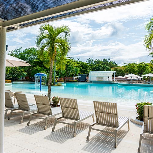 Seis sillas asoleadoras rodeando una piscina que tiene agua cristalina y está iluminada por el sol durante las vacaciones de Lagosol en temporada alta. Alrededor hay parasoles blancos y gran cantidad de vegetación